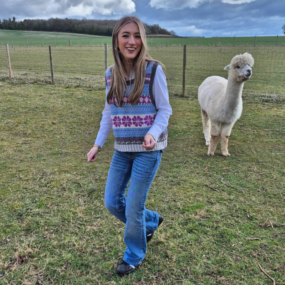 Woman in a colorful sweater and jeans standing next to an alpaca in a field