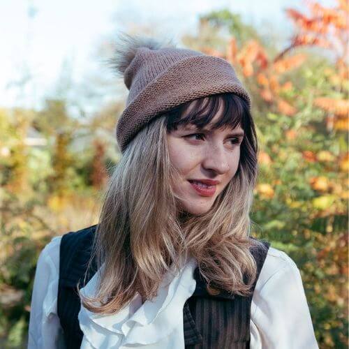Kiki wearing the beginner hat rolled up knitted in DK alpaca wool from British and Irish farms and showing the faux fur pompom